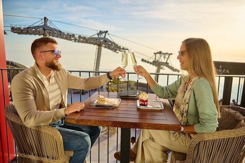 Two people toasting with champagne glasses at a wooden table with a dessert on it, overlooking a scenic view with a bridge in the background. 1