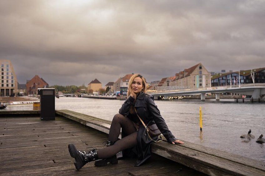 Una persona sentada en un muelle de madera junto a un río, con edificios al fondo. 1