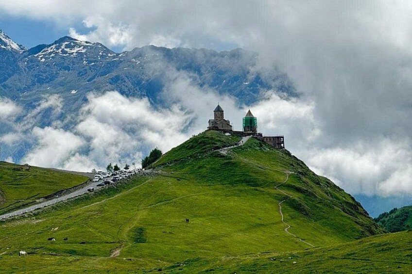 Ein grüner Hügel mit einer Kirche und einem Gebäude auf der Spitze, umgeben von Bergen und Wolken. 3