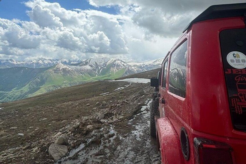 Ein rotes Fahrzeug auf einer Bergstraße, im Hintergrund verschneite Gipfel. 2