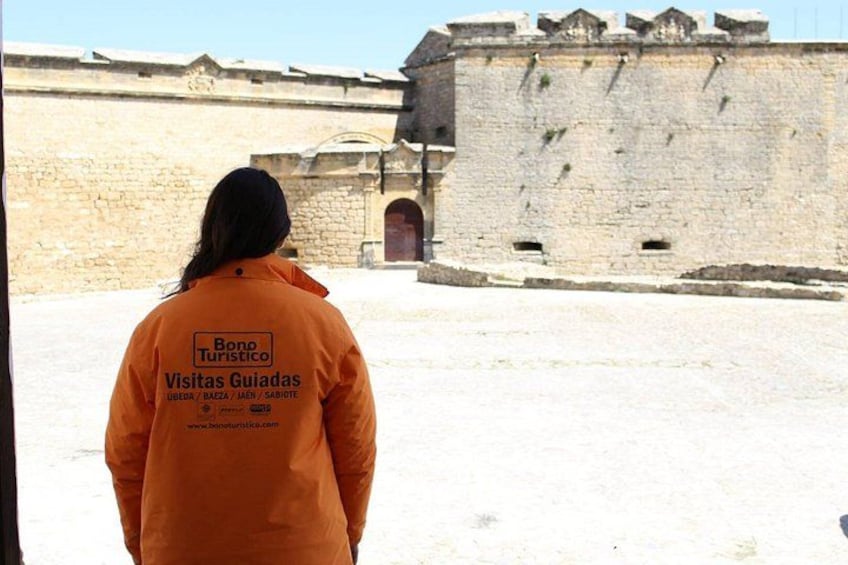 A person in an orange jacket with 'Bono Turismo' text, standing in front of a historical stone wall. 1