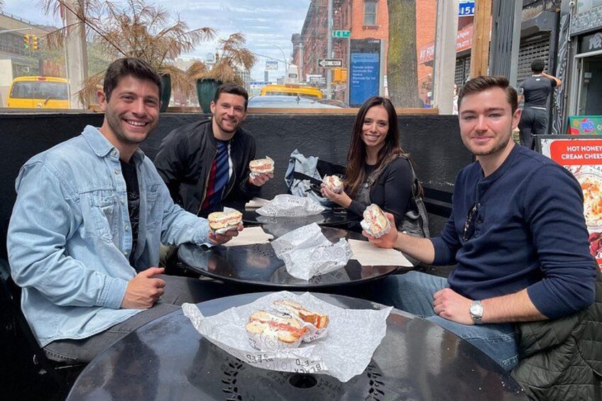 Cuatro personas sentadas en una mesa con comida, una calle de la ciudad al fondo y un letrero que dice 'HOT HONEY CREAM CHEESE'. 4
