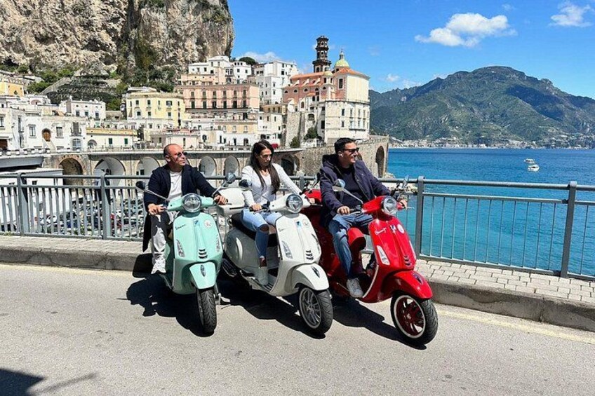 Three people on scooters, a coastal road, and buildings in the background. 5