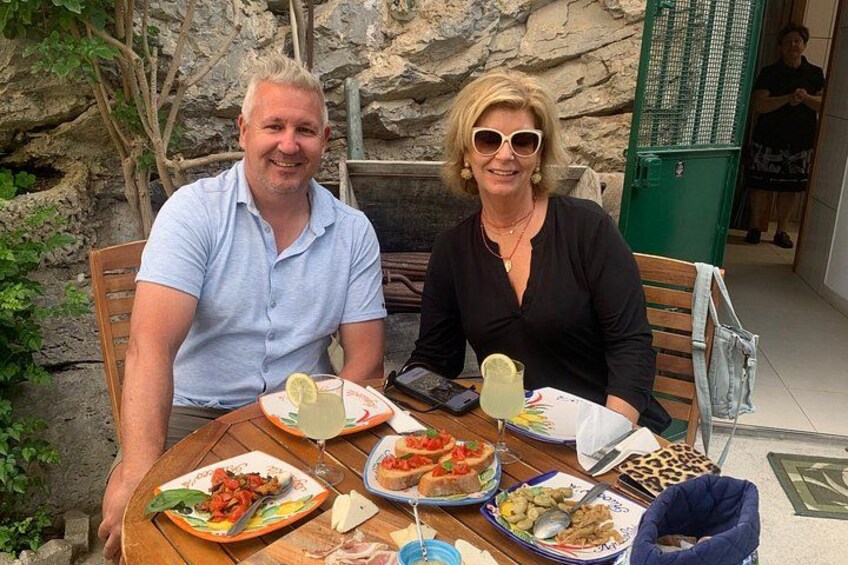 Two people seated at an outdoor table with various dishes, including pasta and a salad, in a rustic setting with stone walls and greenery. 4
