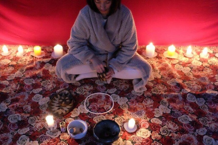A person sitting on a floral carpet with lit candles, surrounded by bowls and a plate. 2