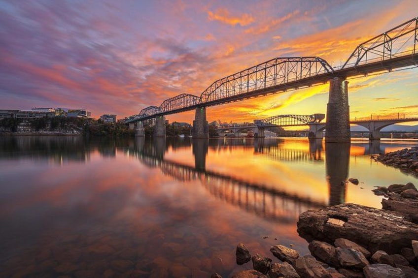 Eine Brücke über einem ruhigen Fluss bei Sonnenuntergang, im Hintergrund eine Stadtansicht. 2