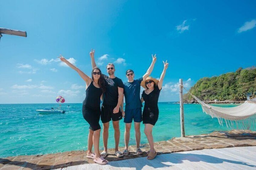 Four people standing on a wooden deck by the sea, with a hammock and a boat in the background. 2