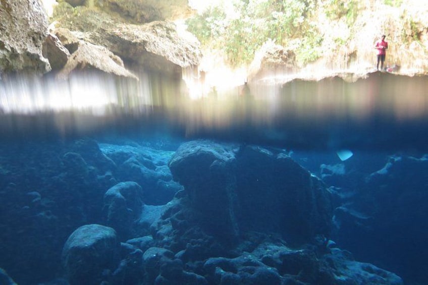 A cave with a person standing on a ledge, a clear water pool, and rocky formations. 4