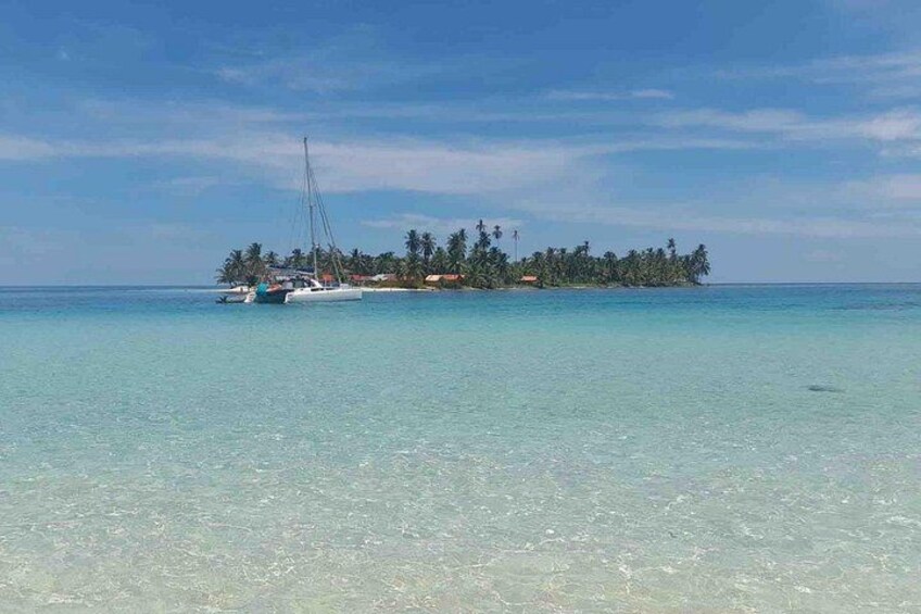 A clear turquoise sea, a small island with palm trees, and a sailboat anchored near the shore. 2