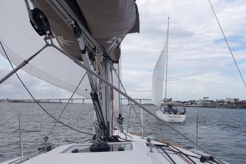 Un velero con vela blanca y una persona en la cubierta, otro velero al fondo, y un puente a lo lejos. 4