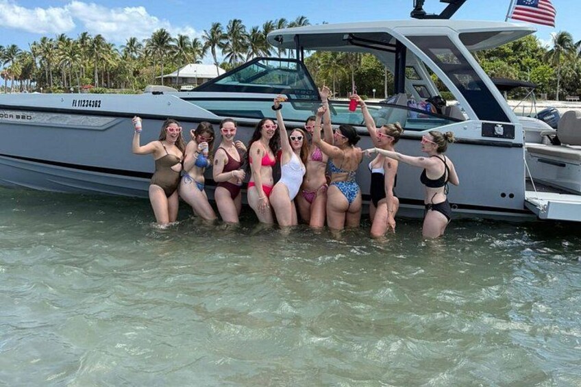 Eine Gruppe von Personen in Badekleidung posiert auf einem Boot auf dem Wasser, im Hintergrund sind Palmen zu sehen. 1