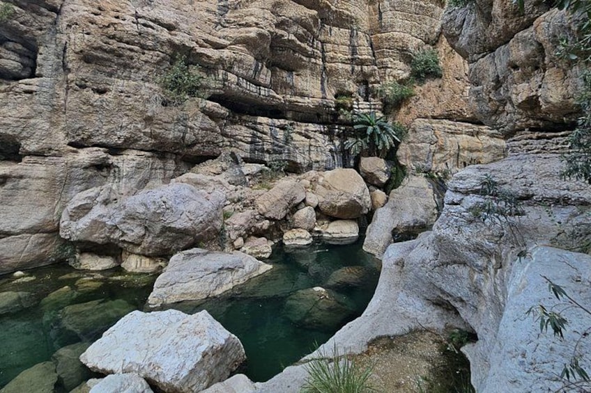 Een rotsachtige kloof met een helder waterbassin, omgeven door grote rotsblokken en schaarse begroeiing. 1