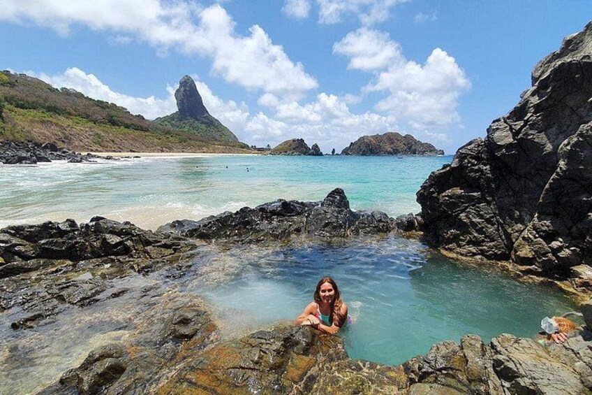 A person in a natural pool surrounded by rocky terrain and a coastal landscape with clear waters and distant mountains. 2