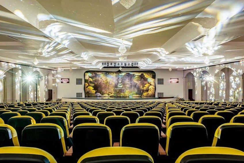 Un teatro con hileras de asientos amarillos, un escenario con una pantalla grande y un techo decorativo. 5