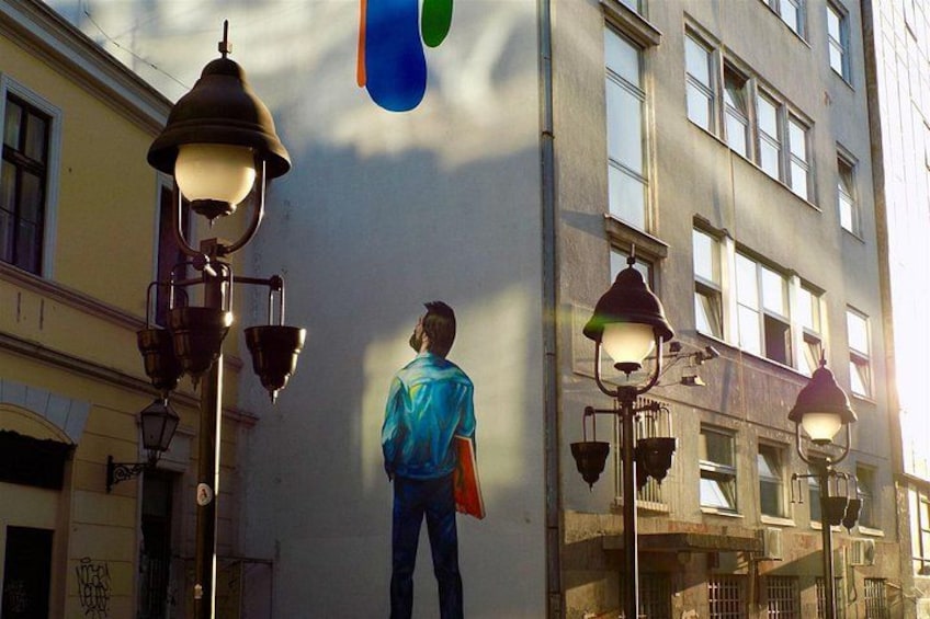 A person standing in front of a building with a mural, street lamps, and a clear sky. 1