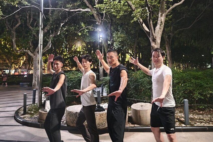 Four people performing a synchronized dance in an outdoor setting with trees and a paved path. 3