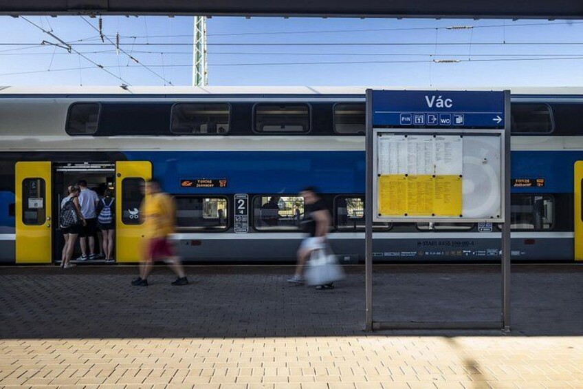 A train station platform with a train, a signboard, and people. 2