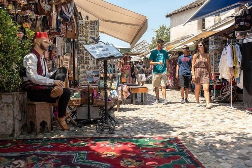 A market scene with a vendor sitting on a stool, a patterned rug on the ground, and various stalls in the background. 3