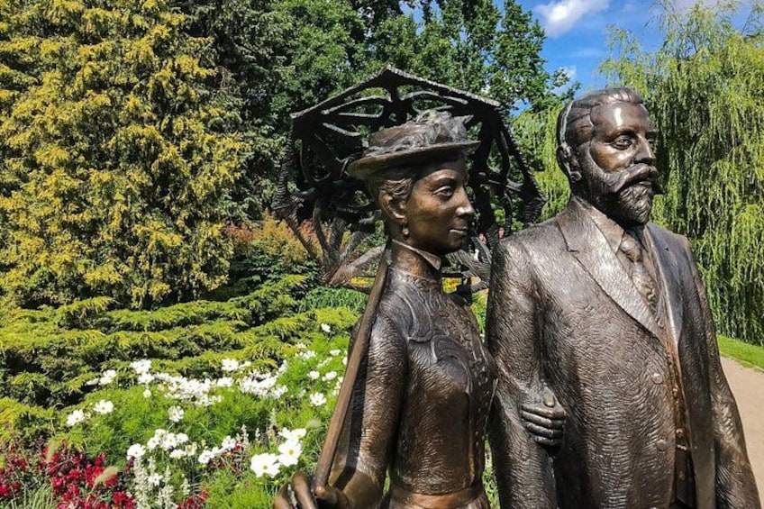 A bronze statue of a man and woman in a park with lush greenery and flowering plants. 1