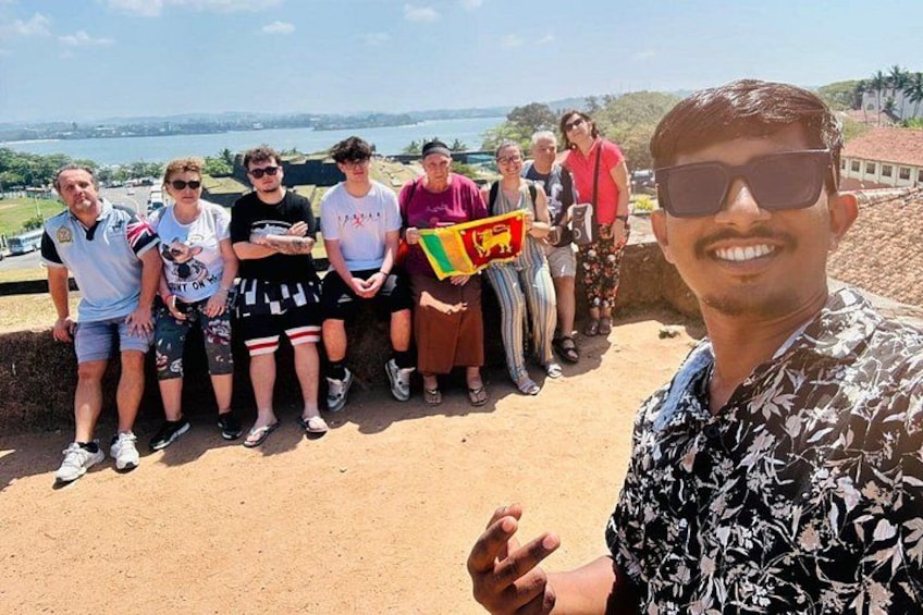 Een groep mensen staat op een zandig gebied, met in de verte een uitzicht op de kust. 1