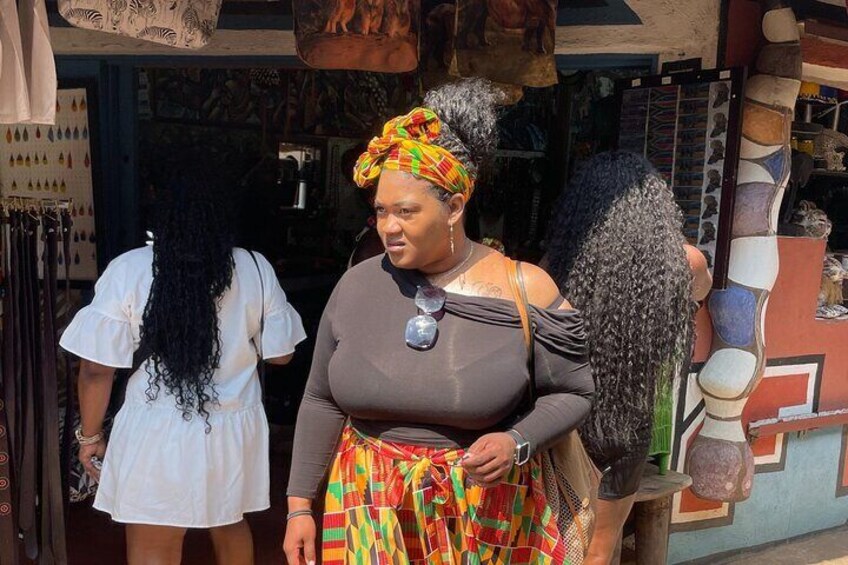 Una persona con un pañuelo colorido y falda estampada, de pie frente a una tienda con diversos artículos expuestos. 5