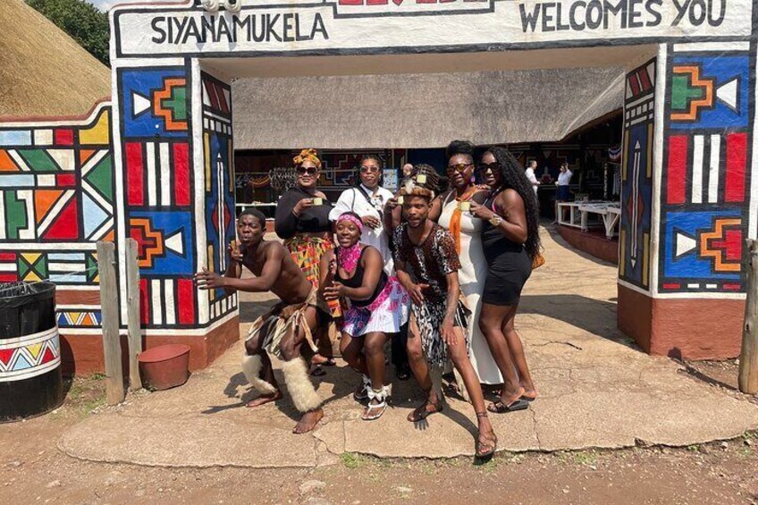 Un grupo de personas posando frente a un letrero colorido que dice 'SIYANAMUKELA WELCOMES YOU'. 3