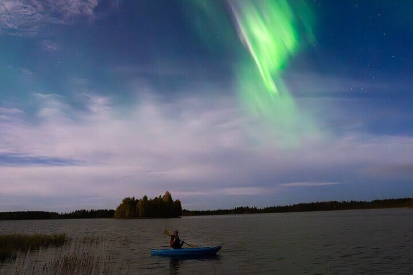 En person som åker kajak på en stilla sjö, med en skogbeklädd ö i bakgrunden och norrsken på himlen. 5