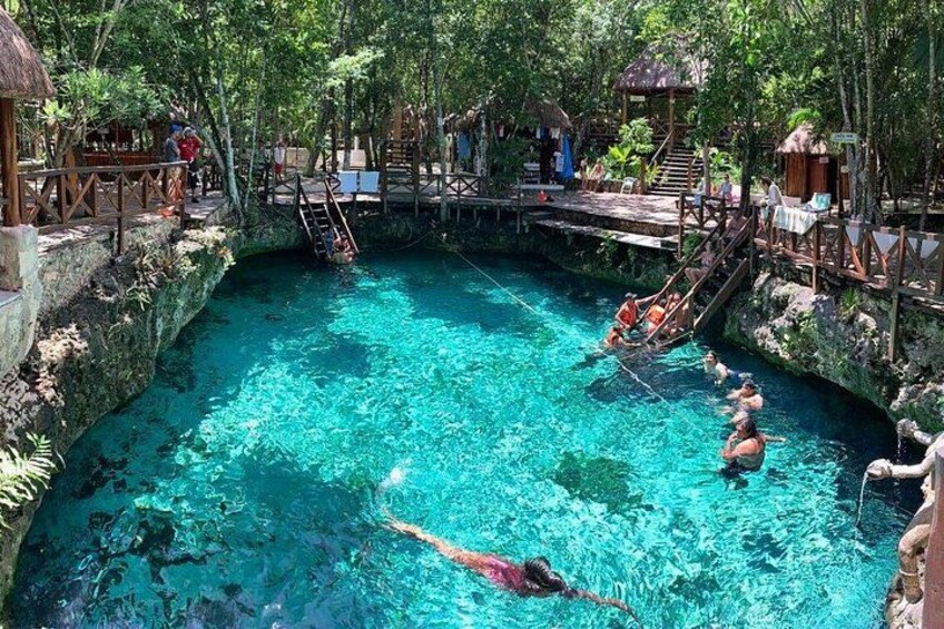 A clear blue pool surrounded by lush greenery, wooden walkways, and a small bridge. 1