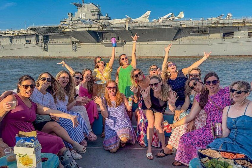 A group of people on a boat with a military aircraft carrier in the background. 1