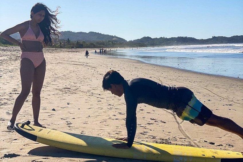 Une personne fait des pompes sur une planche de surf jaune, sur une plage de sable, et une femme en bikini se tient à proximité. 4