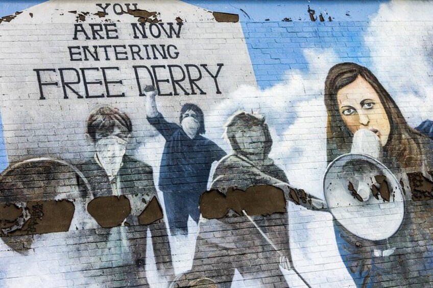 Een muurschildering met mensen die megafoons vasthouden en een bord waarop staat: 'JE BENT NU VRIJE DERRY BINNEN'. 1
