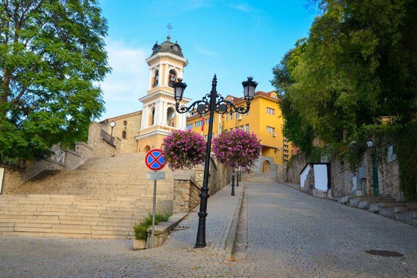 A cobblestone street, a no parking sign, a street lamp, a building with a bell tower, and a flower box with pink flowers. 4