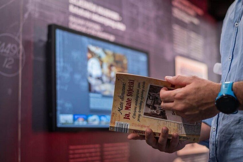 A person holding a book with a visible title and a blurred background with a screen. 3