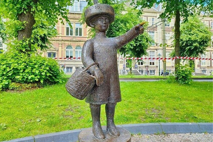 A bronze statue of a child holding a basket, standing on a pedestal in a park with trees and a building in the background. 2