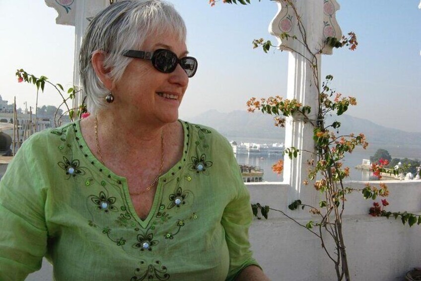 A person wearing a green blouse with floral embroidery, sitting outdoors with a view of mountains and a white structure with flowers. 4