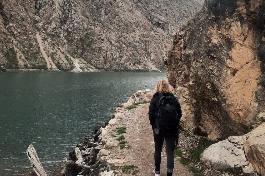 A person hiking along a rocky path beside a lake with steep cliffs in the background. 1