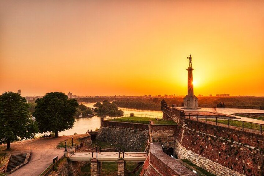A sunset over a historic fort with a statue, a river, and a park. 2
