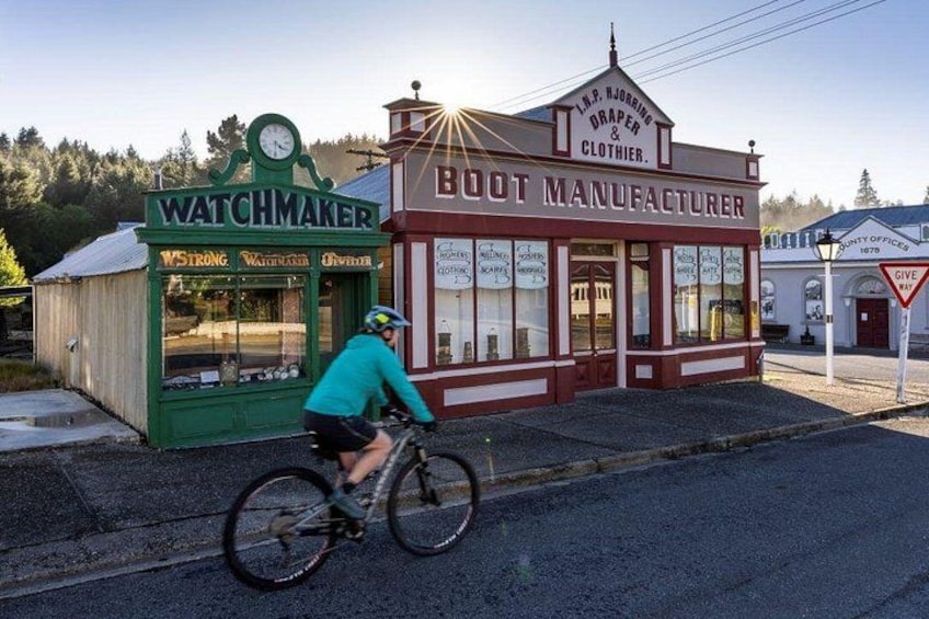 A cyclist riding past a green storefront labeled 'WATCHMAKER' and a red building labeled 'BOOT MANUFACTURER.' 1