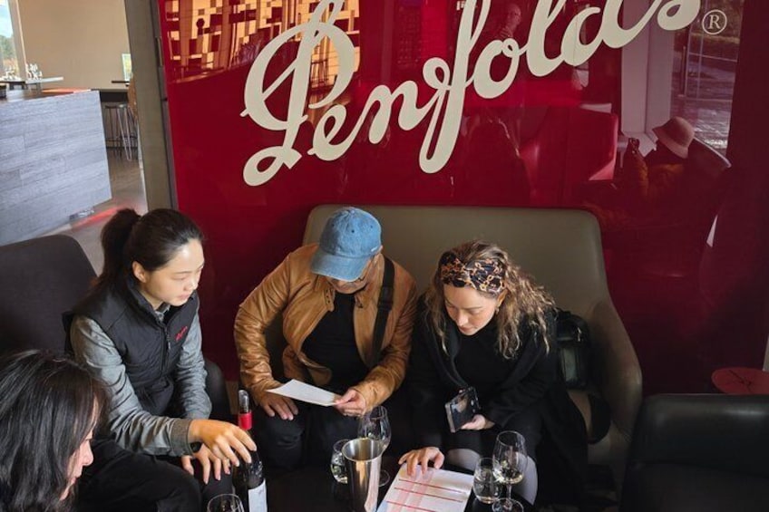 Drei Personen sitzen an einem Tisch mit Weingläsern und einer Speisekarte in einem Restaurant mit roter Wand und einem großen 'Penfolds'-Schild. 2