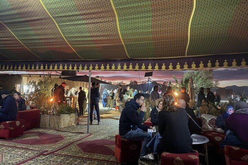 An outdoor gathering with people seated on red cushioned chairs under a patterned canopy. 5