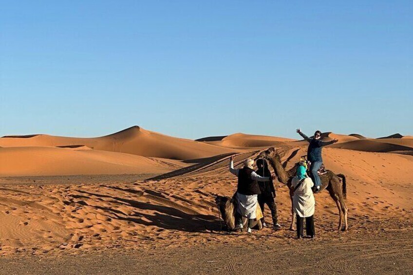 A group of people in a desert with sand dunes, some standing and others riding camels. 1