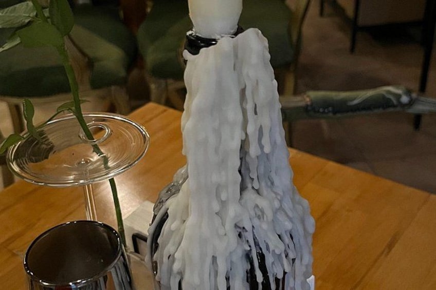 Ein weißer Keramik-Hahn mit weißem Zuckerguss auf dem Rücken, eine transparente Glasvase mit einer grünen Pflanze und eine Metalltasse auf einem Holztisch. 2