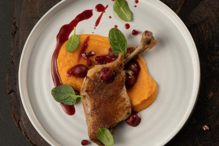 A plate with a roasted duck leg, a dollop of orange puree, and red sauce, garnished with green leaves. 4