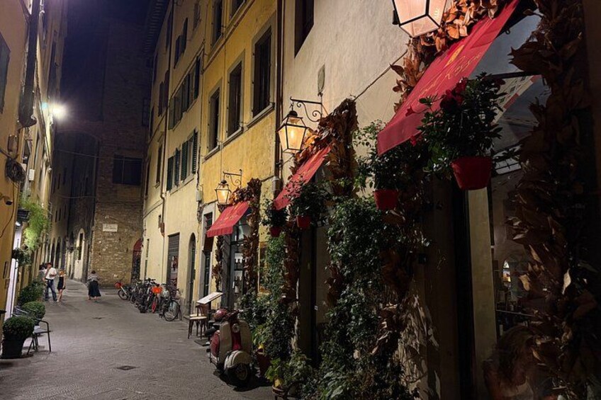 Une rue étroite, la nuit, bordée de bâtiments des deux côtés, dont certains sont dotés d’auvents rouges et de plantes. 5