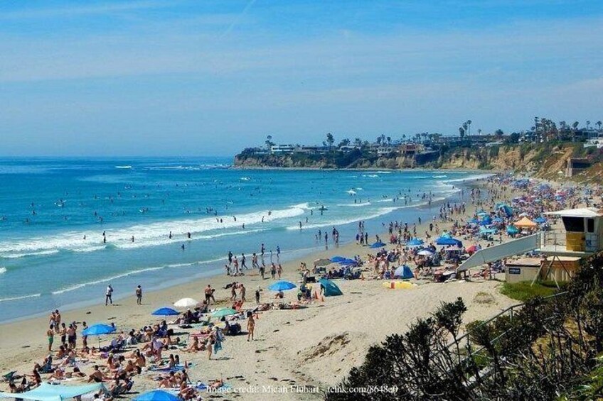 Una playa concurrida, gente tomando el sol y nadando, una torre de salvavidas y un litoral rocoso. 4