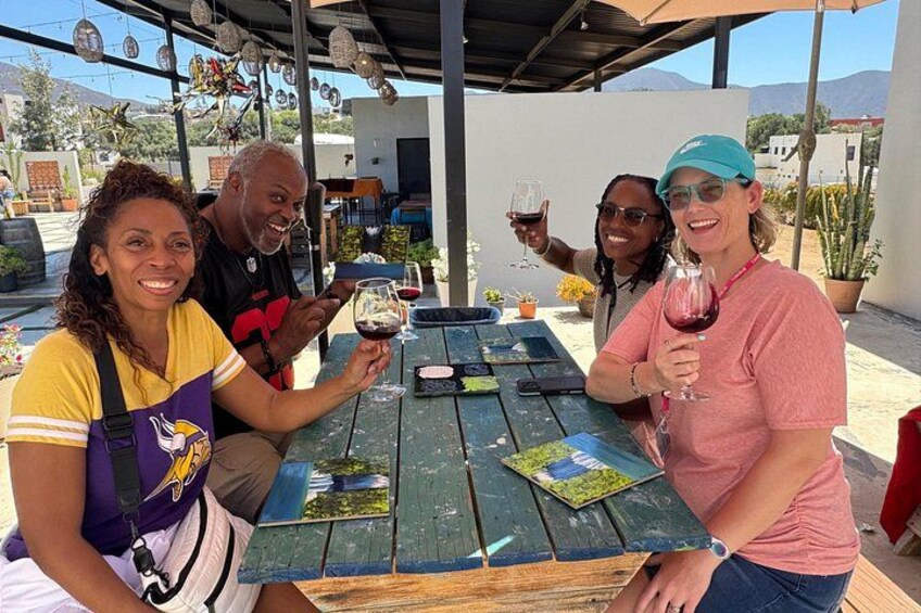 A group of people sitting at an outdoor table with drinks, a wooden table, and a covered area with plants and a mountain in the background. 3