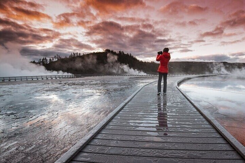 A person in a red jacket taking a photo on a wooden boardwalk by a geothermal area with steam rising from the ground. 3