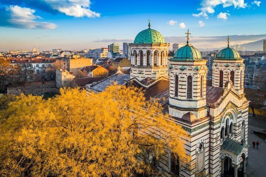 A historic church with green domes and a bell tower, surrounded by autumn trees and urban buildings. 1
