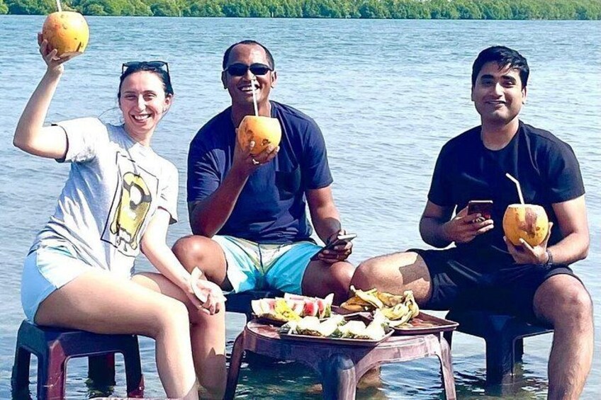 Tre persone sedute su una barca, con delle noci di cocco in mano, con uno specchio d'acqua e vegetazione sullo sfondo. 3