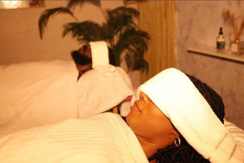 A person lying in bed with a white towel over their head, a potted plant, and a shelf with items in the background. 1